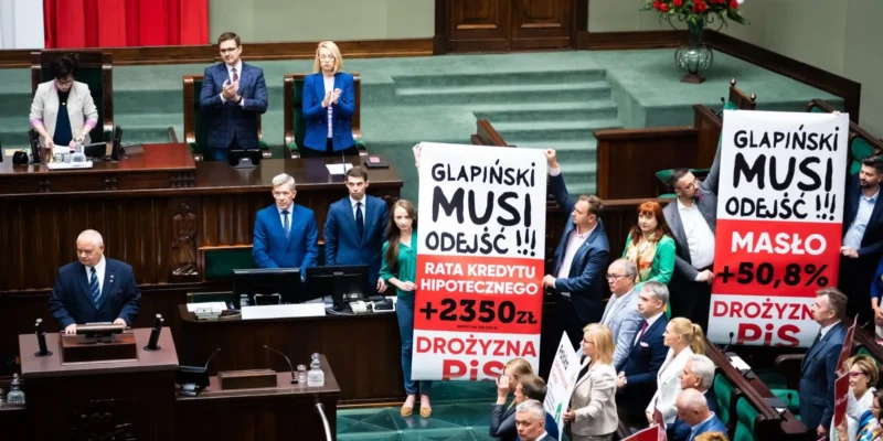 Na zdjęciu odbywa się protest w polskim parlamencie, w czasie którego demonstranci trzymają transparenty z hasłami dotyczącymi dymisji Glapińskiego oraz wzrostu kosztów życia.