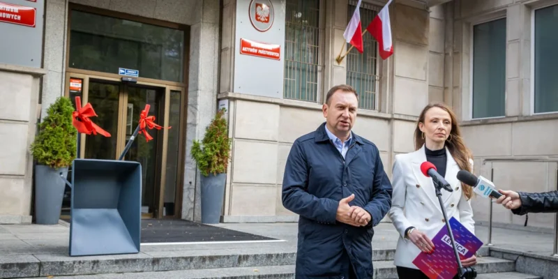 Na zdjęciu widać dwoje ludzi stojących przed budynkiem, jeden z nich trzyma mikrofon, a obok nich znajduje się łopata ozdobiona czerwonymi wstążkami.