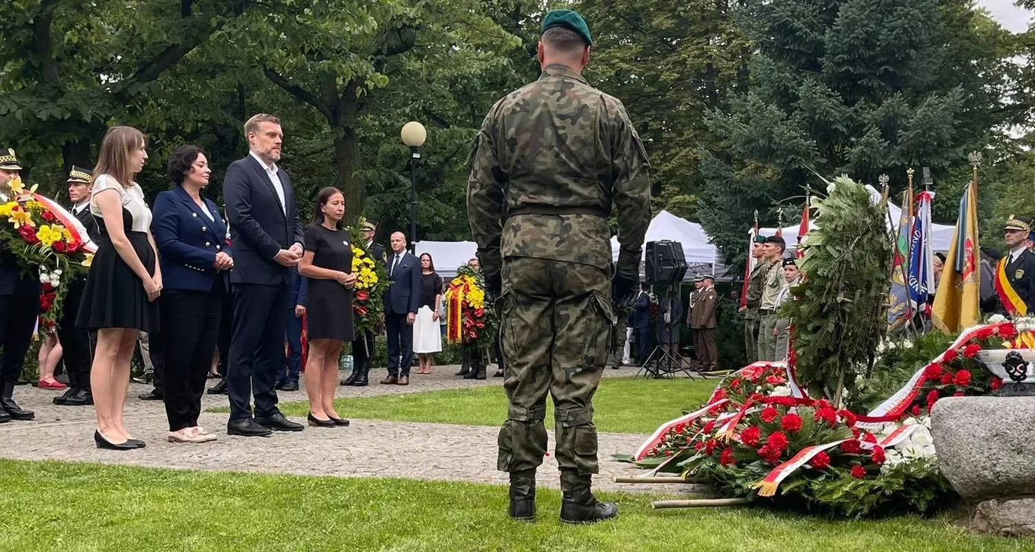 Grupa osób w mundurach i garniturach składa hołd bohaterom i ofiarom Powstania Warszawskiego, stojąc obok wiązanek kwiatów i flag w parku.