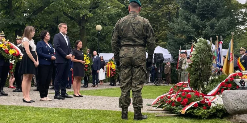 Grupa osób w mundurach i garniturach składa hołd bohaterom i ofiarom Powstania Warszawskiego, stojąc obok wiązanek kwiatów i flag w parku.