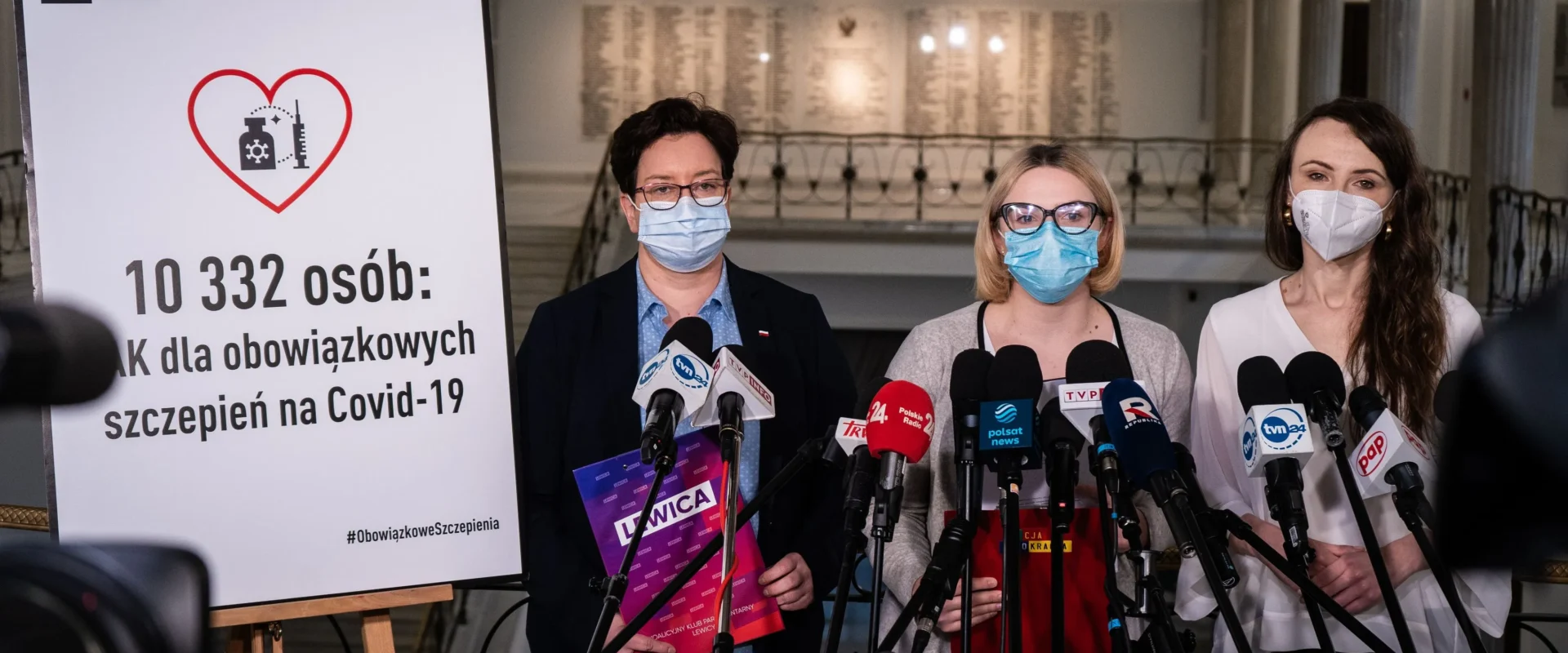 Na zdjęciu przedstawione są trzy kobiety w maseczkach, które stoją przed banerem promującym obowiązkowe szczepienia na COVID-19, podczas konferencji prasowej.