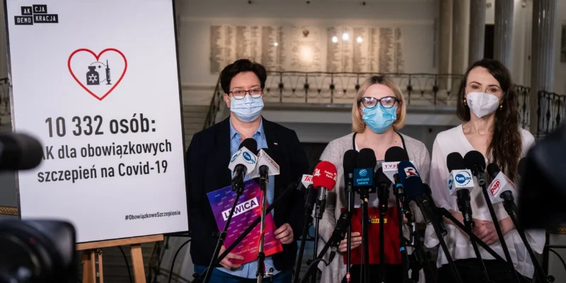 Na zdjęciu przedstawione są trzy kobiety w maseczkach, które stoją przed banerem promującym obowiązkowe szczepienia na COVID-19, podczas konferencji prasowej.