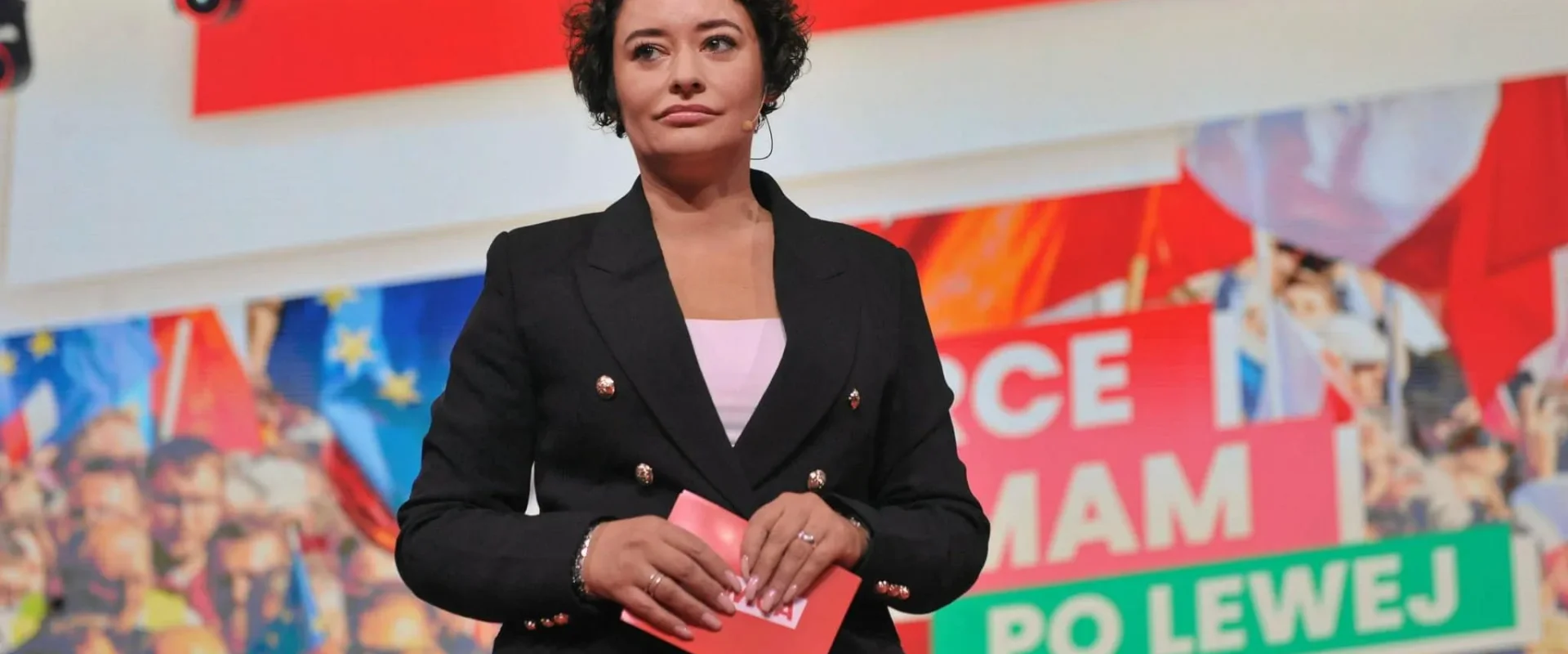 Polityczka w czarnym garniturze przemawia na scenie z mikroportem podczas wydarzenia o dopłatach do kredytów, z kolorowymi banerami i tłumem w tle.