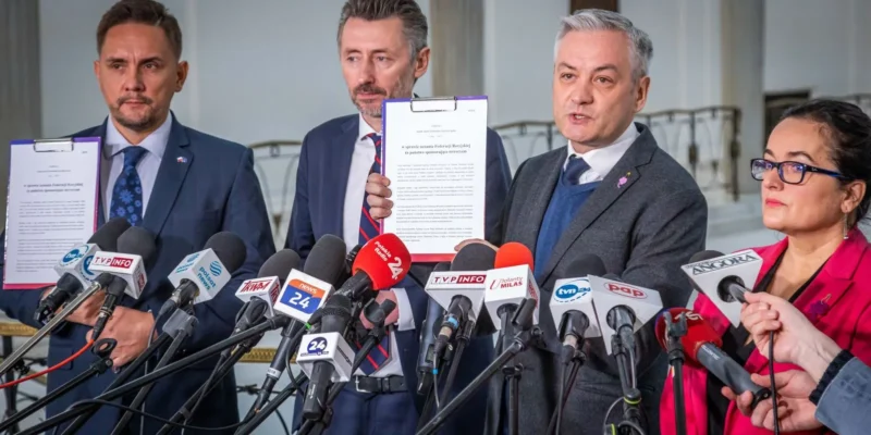 Na zdjęciu przedstawiono czterech polityków podczas konferencji prasowej, na której prezentują dokument i odpowiadają na pytania dziennikarzy, otoczeni mikrofonami mediów.