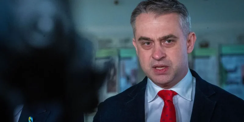 Mężczyzna w garniturze z czerwonym krawatem przemawia do mediów, wyrażając emocje w poważnej atmosferze.