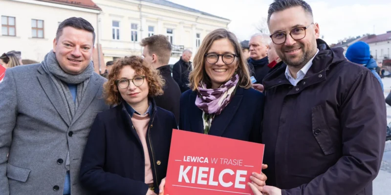 Czwórka uśmiechniętych osób stoi na placu w Kielcach, trzymając czerwony plakat z napisem „LEWICA W TRASIE KIELCE” podczas wydarzenia w województwie świętokrzyskim.