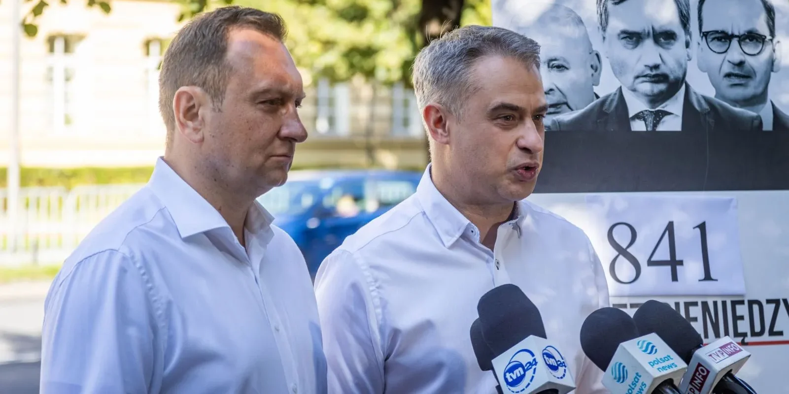 Dwóch mężczyzn w białych koszulach udziela konferencji prasowej przed mikrofonami, w tle widoczny plakat z twarzami, w kontekście incydentu z udziałem Black Hawk.