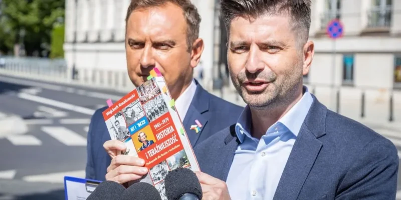 Na zdjęciu dwóch mężczyzn, jeden z nich trzyma książkę i mówi, podczas gdy drugi stoi w tle, na tle miejskiego krajobrazu.