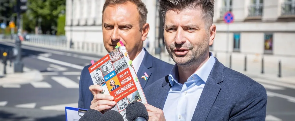 Na zdjęciu dwóch mężczyzn, jeden z nich trzyma książkę i mówi, podczas gdy drugi stoi w tle, na tle miejskiego krajobrazu.