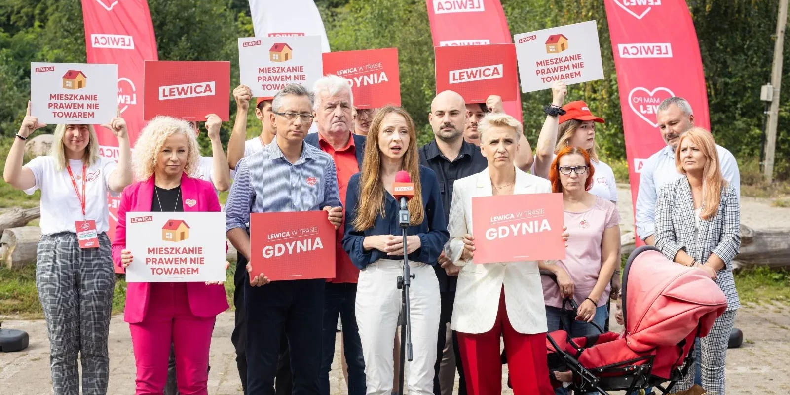 Grupa zwolenników Lewicy na zewnątrz trzyma plakaty z hasłami „MIESZKANIE PRAWEM NIE TOWAREM” i „GDYNIA”, stojąc wokół mikrofonu, z czerwonymi flagami Lewicy w tle.