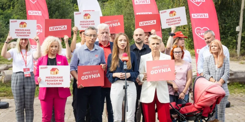 Grupa zwolenników Lewicy na zewnątrz trzyma plakaty z hasłami „MIESZKANIE PRAWEM NIE TOWAREM” i „GDYNIA”, stojąc wokół mikrofonu, z czerwonymi flagami Lewicy w tle.