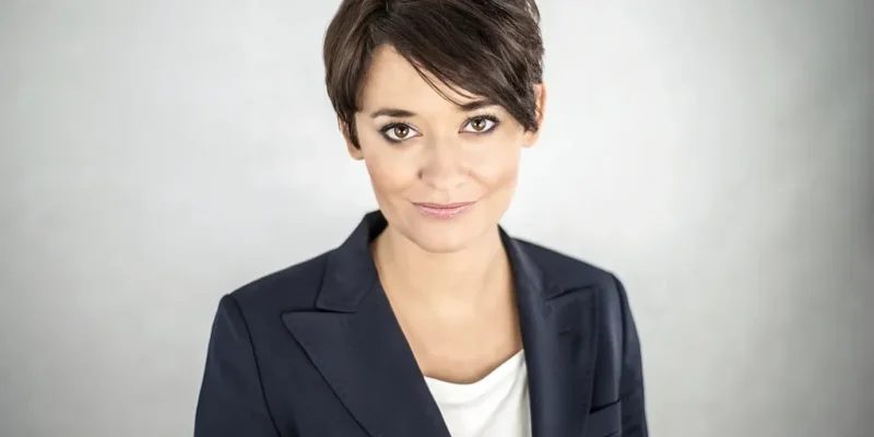 Portret kobiety o krótkich ciemnych włosach, ubranej w granatową marynarkę i białą bluzkę, patrzącej prosto w obiektyw na jasnoszarym tle.