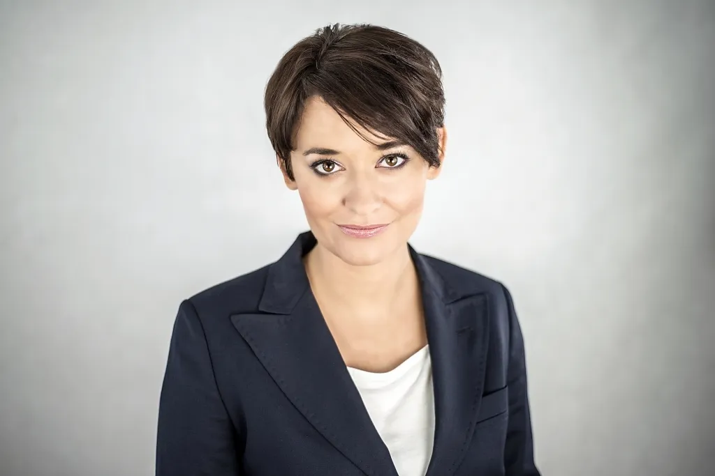 Portret kobiety o krótkich ciemnych włosach, ubranej w granatową marynarkę i białą bluzkę, patrzącej prosto w obiektyw na jasnoszarym tle.