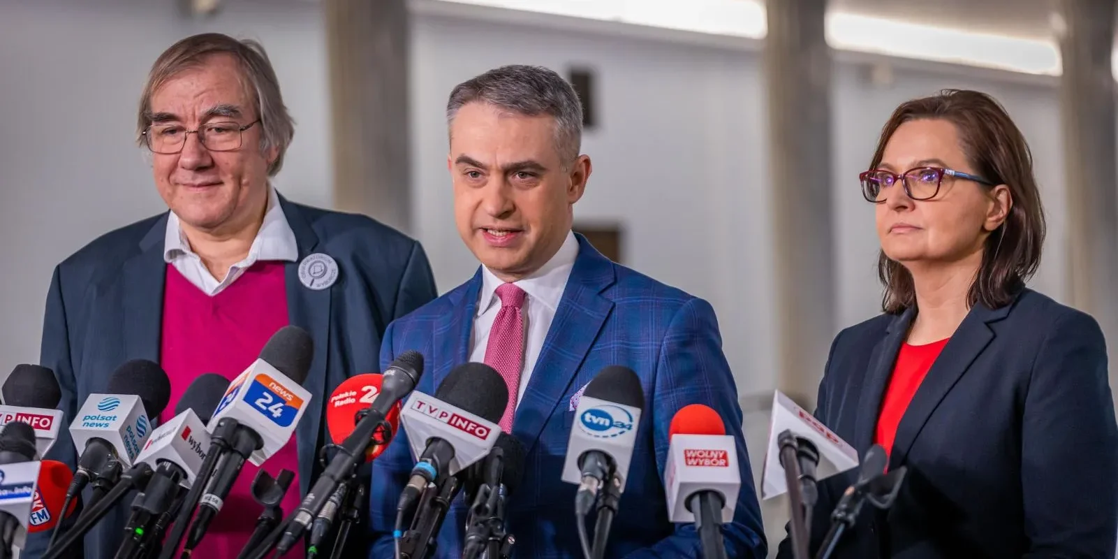 Trzech polityków stoi na konferencji prasowej otoczeni licznymi mikrofonami mediów, omawiając temat Kodeksu Wyborczego.