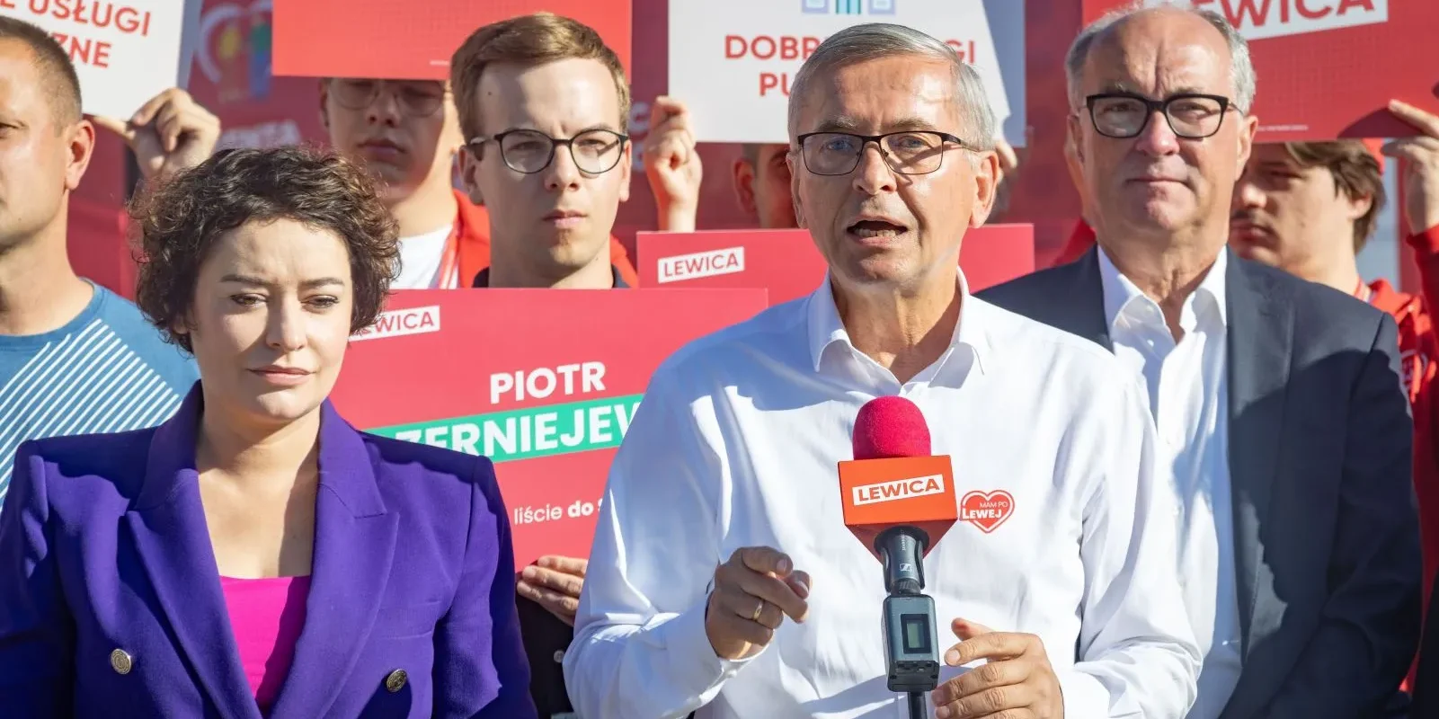 Grupa osób na konferencji Lewicy; na pierwszym planie mężczyzna w białej koszuli przemawia do mikrofonu z logiem Lewicy, a obok niego stoi kobieta w fioletowej marynarce, w tle widoczne plakaty Lewicy.