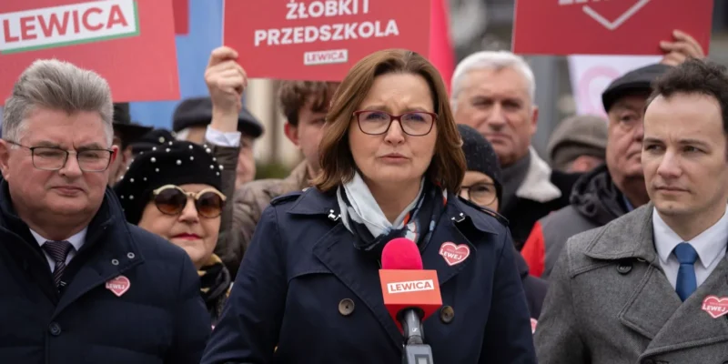 Kobieta w okularach z czerwonymi oprawkami przemawia do zgromadzonej publiczności podczas wiecu Lewicy, trzymając różowy mikrofon na tle plakatów.