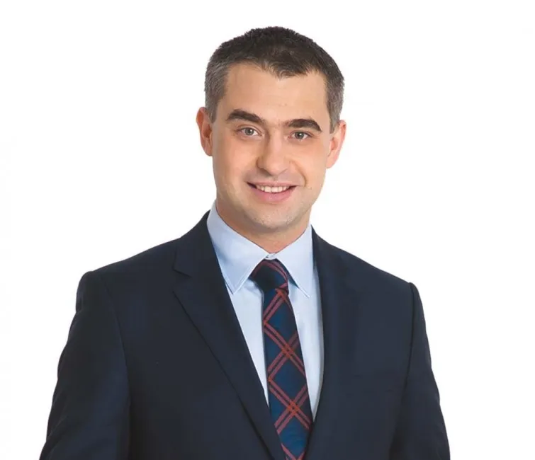 Uśmiechnięty mężczyzna w granatowym garniturze i kraciastym krawacie na białym tle.