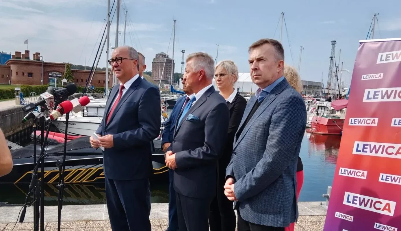 Na zdjęciu widoczni są mężczyźni i kobieta stojący przy porcie, omawiający sprawy polityczne, w tle łodzie i baner z napisem "LEWICA".