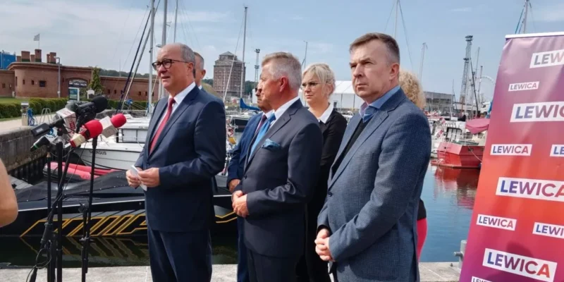 Na zdjęciu widoczni są mężczyźni i kobieta stojący przy porcie, omawiający sprawy polityczne, w tle łodzie i baner z napisem "LEWICA".