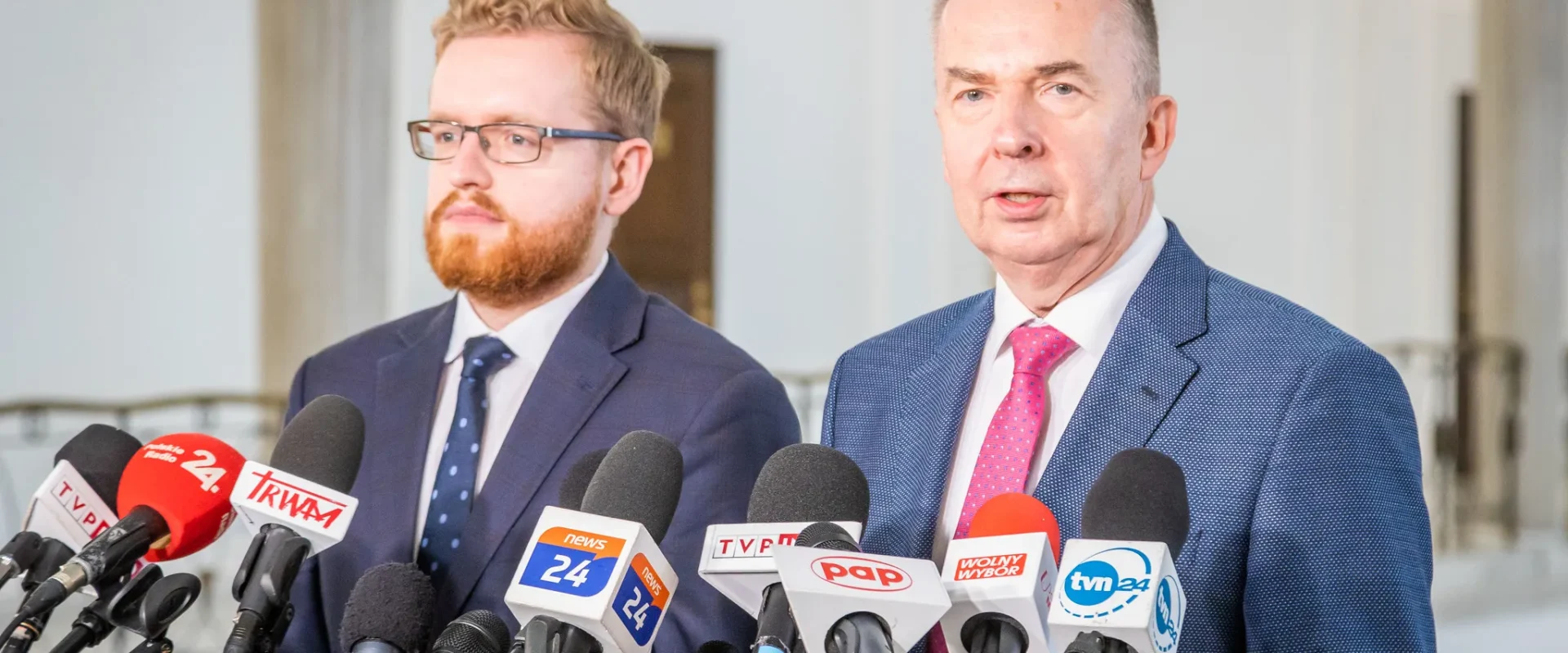 Dwóch mężczyzn w garniturach stoi przed mikrofonami podczas konferencji prasowej w jasnym wnętrzu.