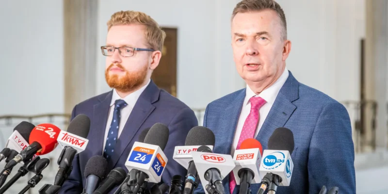 Dwóch mężczyzn w garniturach stoi przed mikrofonami podczas konferencji prasowej w jasnym wnętrzu.