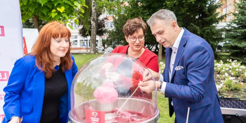 Trzy osoby stoją w parku przy maszynie do waty cukrowej podczas wydarzenia Trasa Bezpieczna Rodzina.