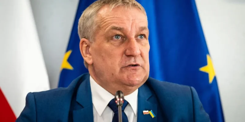 Mężczyzna w granatowym garniturze przemawia podczas konferencji, w tle widoczne flagi Polski i Unii Europejskiej.