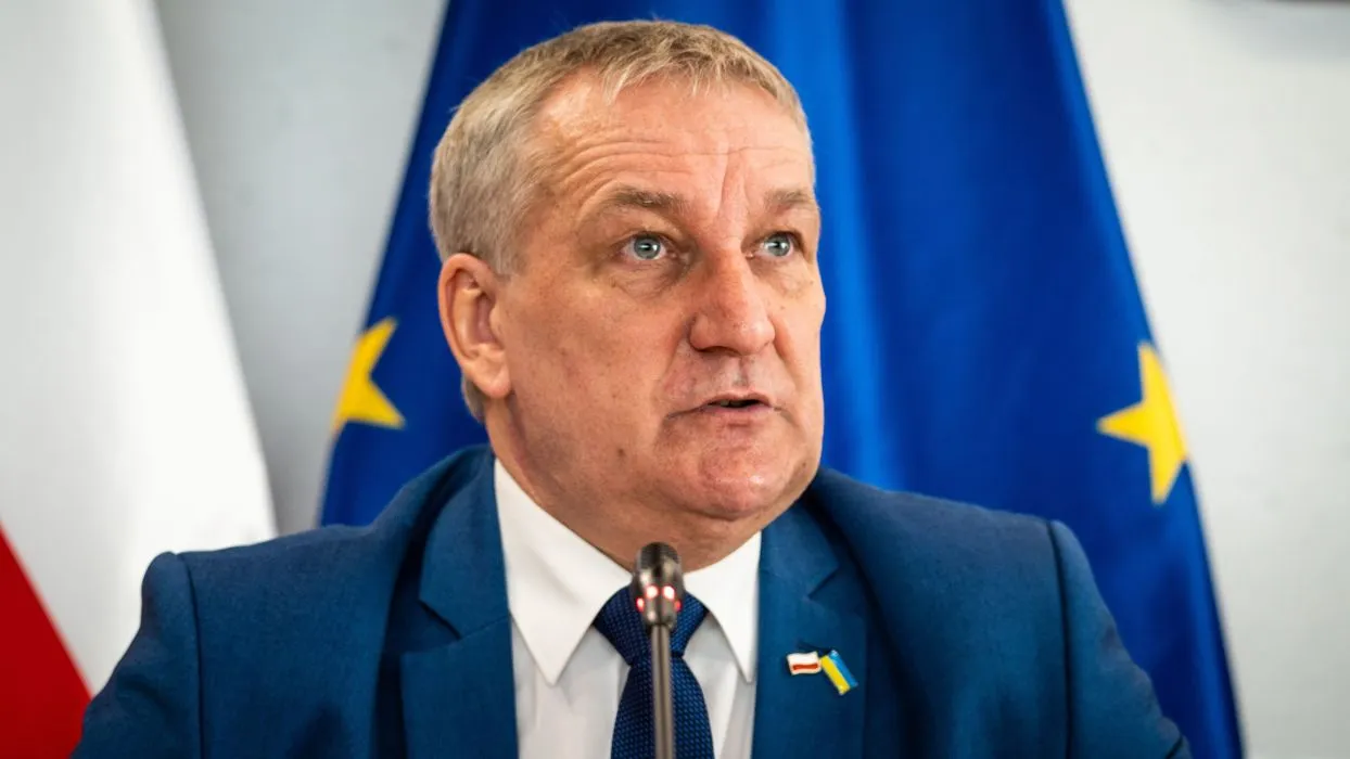 Mężczyzna w granatowym garniturze przemawia podczas konferencji, w tle widoczne flagi Polski i Unii Europejskiej.