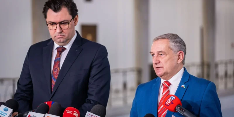 Na zdjęciu dwóch mężczyzn w eleganckich garniturach przemawia do mediów, podczas gdy w tle widać mikrofony z logo różnych stacji informacyjnych.