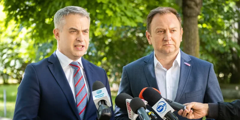 Dwóch mężczyzn w garniturach prowadzi konferencję prasową w zielonym parku, otoczeni mikrofonami dziennikarzy.