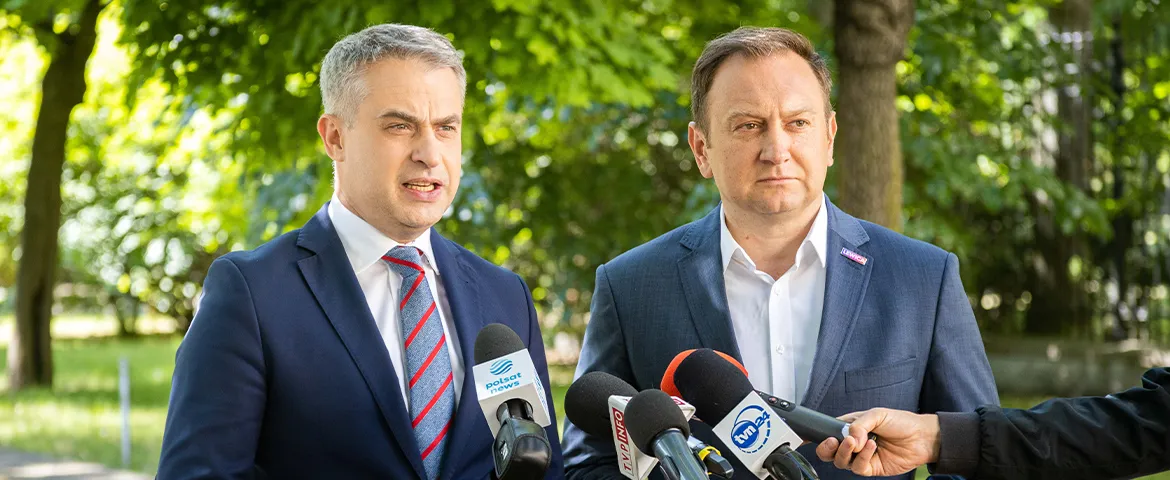 Dwóch mężczyzn w garniturach prowadzi konferencję prasową w zielonym parku, otoczeni mikrofonami dziennikarzy.