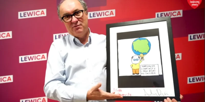 Mężczyzna w okularach trzyma w ramce rysunek przedstawiający Ziemię nad żółtą postacią, na czerwonym tle z logami Lewicy, podczas wydarzenia Wielkiej Orkiestry Świątecznej Pomocy.