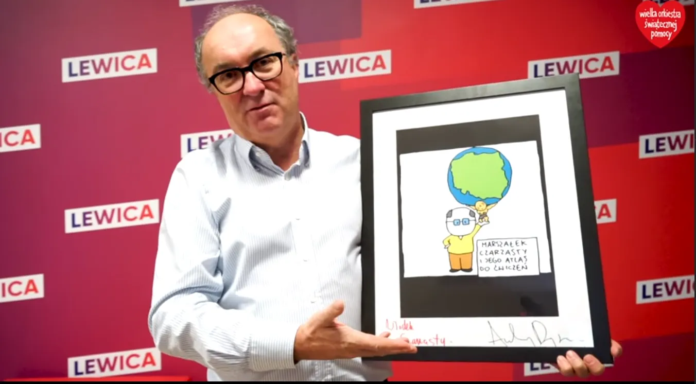 Mężczyzna w okularach trzyma w ramce rysunek przedstawiający Ziemię nad żółtą postacią, na czerwonym tle z logami Lewicy, podczas wydarzenia Wielkiej Orkiestry Świątecznej Pomocy.