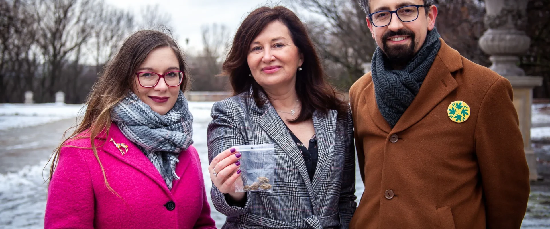 Na zdjęciu widać trzy osoby stojące na zewnątrz w zimowej scenerii, z jedną z nich trzymającą woreczek z materiałem, w otoczeniu nagich drzew i śniegu.