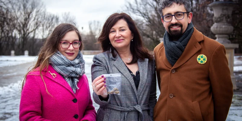Na zdjęciu widać trzy osoby stojące na zewnątrz w zimowej scenerii, z jedną z nich trzymającą woreczek z materiałem, w otoczeniu nagich drzew i śniegu.