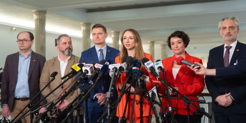 Grupa polityków stoi na konferencji prasowej, otoczona licznymi mikrofonami, omawiając kwestię renty wdowiej.