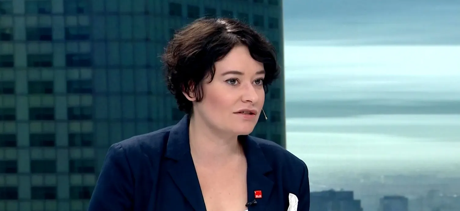 Kobieta w granatowym żakiecie z mikrofonem na klipsie rozmawia w studiu telewizyjnym, a w tle widać panoramę miasta.