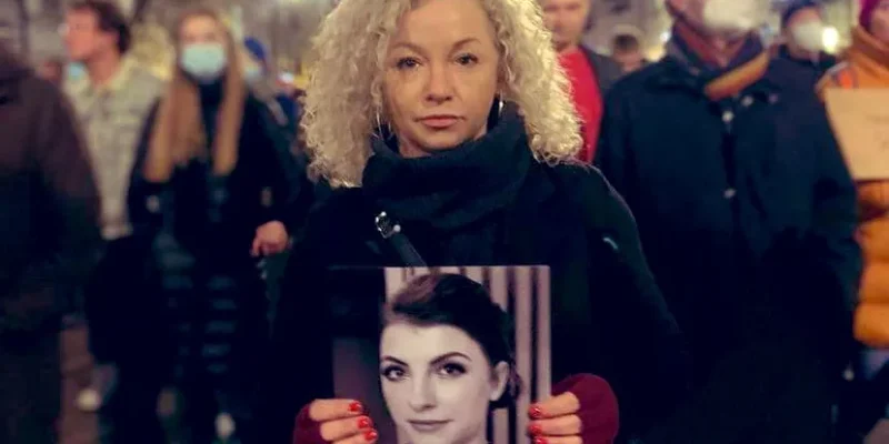 Protestująca kobieta stoi w tłumie na nocnej ulicy, trzyma plakat z czarno-białym portretem.