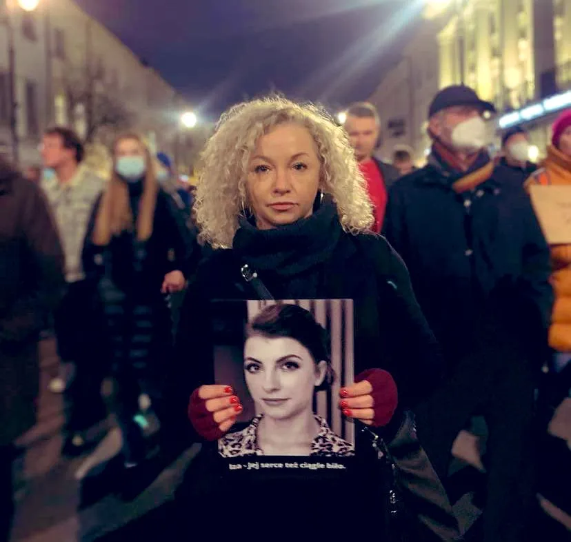 Protestująca kobieta stoi w tłumie na nocnej ulicy, trzyma plakat z czarno-białym portretem.