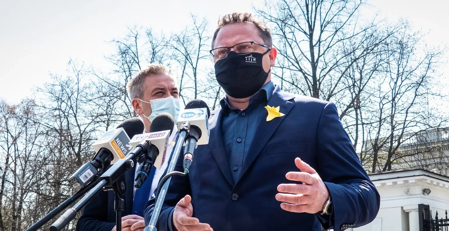 Dwóch mężczyzn w garniturach podczas konferencji prasowej stojących przy mikrofonach; jeden w czarnej masce z żółtą ozdobą na klapie, w tle bezlistne drzewa.