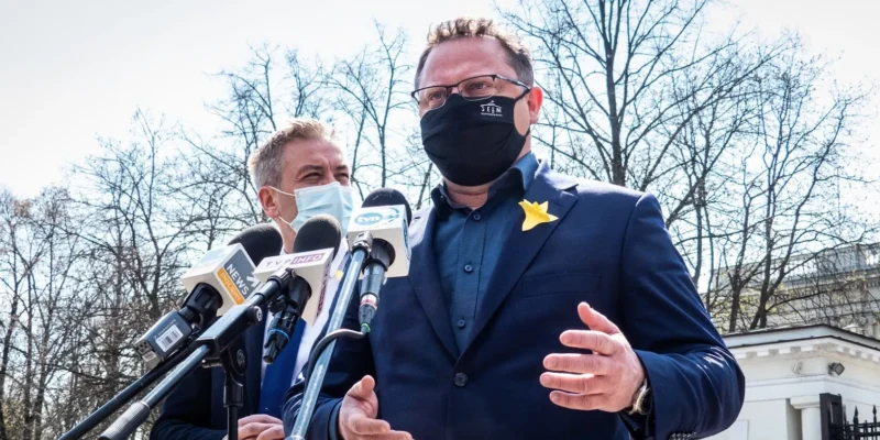 Dwóch mężczyzn w garniturach podczas konferencji prasowej stojących przy mikrofonach; jeden w czarnej masce z żółtą ozdobą na klapie, w tle bezlistne drzewa.