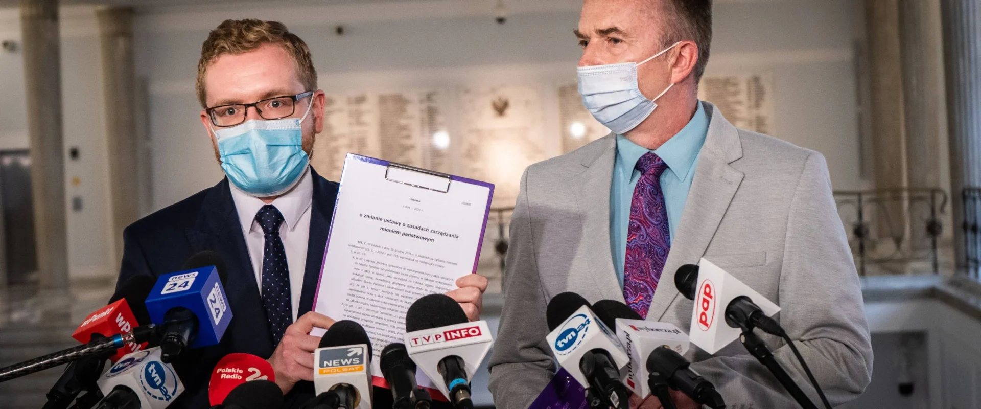 Dwóch mężczyzn w garniturach i maseczkach na konferencji prasowej, otoczonych licznymi mikrofonami, z jednym trzymającym kartkę z dokumentem.