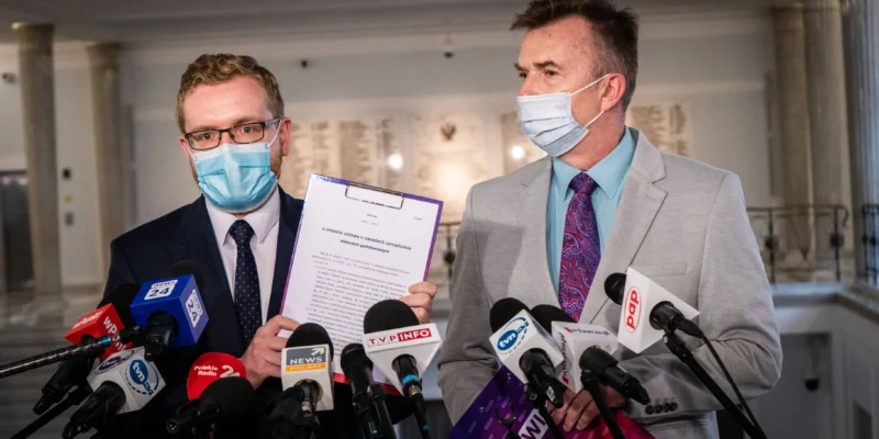 Dwóch mężczyzn w garniturach i maseczkach na konferencji prasowej, otoczonych licznymi mikrofonami, z jednym trzymającym kartkę z dokumentem.