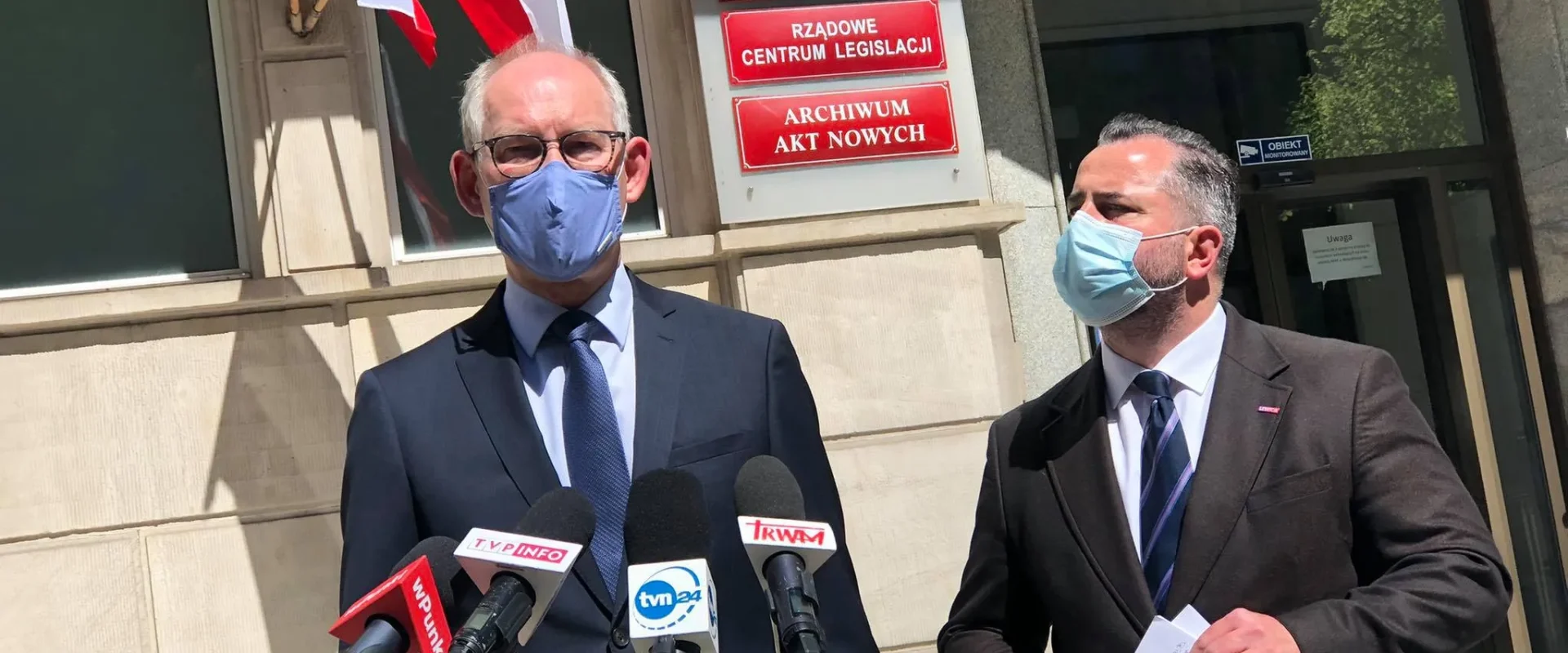 Dwóch mężczyzn w garniturach i maseczkach stoi przed zestawem mikrofonów podczas konferencji prasowej, obok flagi Polski i wejścia do budynku rządowego.