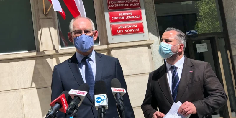 Dwóch mężczyzn w garniturach i maseczkach stoi przed zestawem mikrofonów podczas konferencji prasowej, obok flagi Polski i wejścia do budynku rządowego.