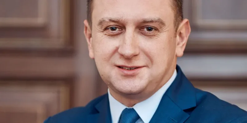 Półzbliżeniowy portret uśmiechniętego mężczyzny w granatowym garniturze i niebieskim krawacie na tle zdobionych drewnianych drzwi.