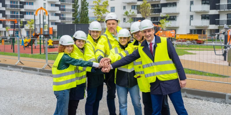 Grupa osób w odblaskowych kamizelkach i kaskach budowlanych wspólnie podnosi ręce, symbolizując współpracę na tle placu budowy z nowym placem zabaw.