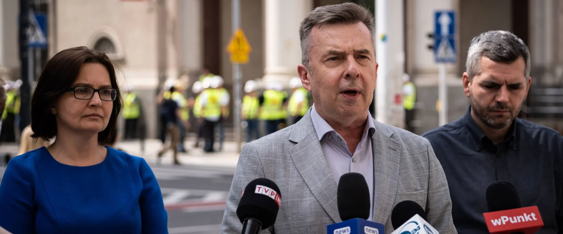 Mężczyzna w jasnoszarym garniturze przemawia do mediów podczas konferencji prasowej na ulicy, otoczony przez współpracowników i liczne mikrofony z logami stacji telewizyjnych.