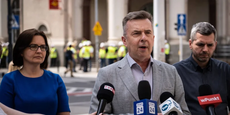 Mężczyzna w jasnoszarym garniturze przemawia do mediów podczas konferencji prasowej na ulicy, otoczony przez współpracowników i liczne mikrofony z logami stacji telewizyjnych.