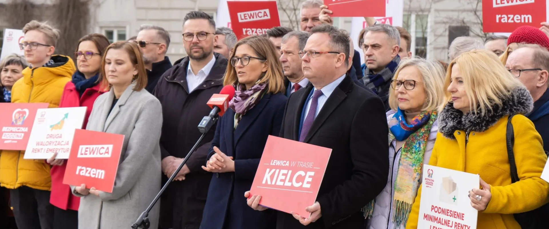 Na zdjęciu widoczna jest grupa osób biorących udział w konferencji prasowej, trzymających transparenty z hasłami politycznymi, w tym „LEWICA w trasie KIELCE” i „LEWICA razem”.
